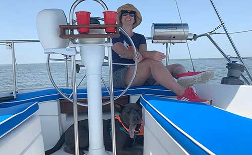 A smiling woman sits behind the wheel of a sailboat on a lake while a large black dog with big ears lies on the deck below her. 